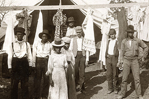 Men and women look straight on as they stand outside of a tent and a clothesline.