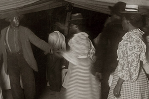 A man and woman hold hands as they dance. Others look on and some look towards the camera.