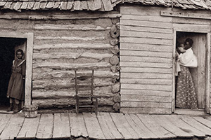 A woman holds a baby and a young girl stares straight on in the doorways of a log cabin.