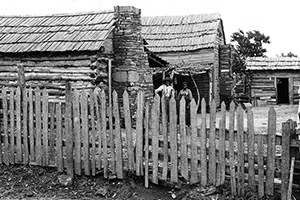 Men and women stand outside their log cabins, which are enclosed by a picket fence.
