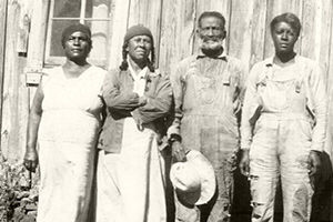 An older man stands outside a wooden structure with three women