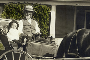 An older man sits next to small white child in a horse-drawn buggy.