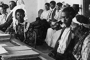 Several men and women sit in a large room behind a wooden bar. A few individuals sit at large desks on the other side.
