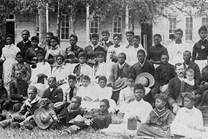 A large group of men, women, and children sit in rows in front of the school.