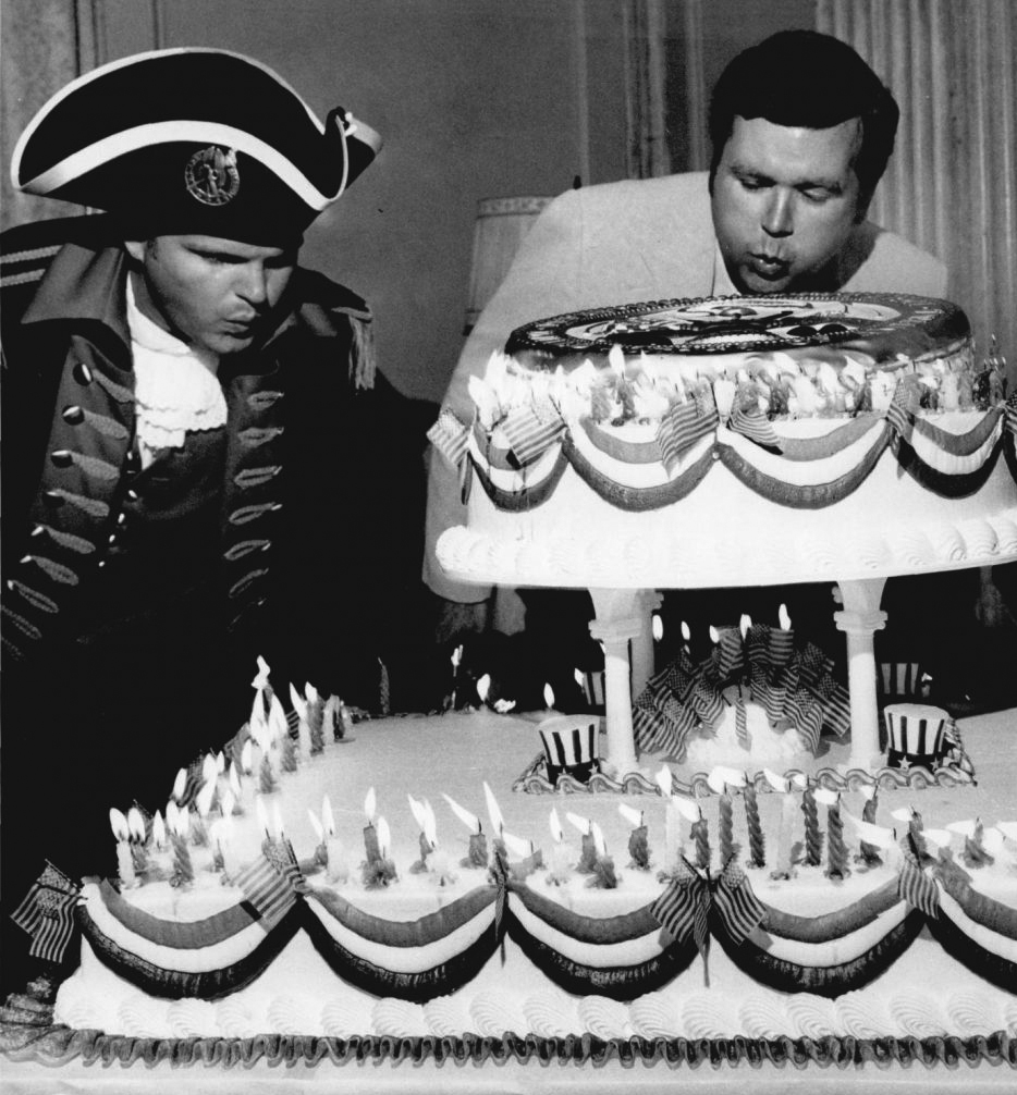Governor Boren and a man dressed in a Revolutionary War uniform lean forward to blow out candles on a large cake. The cake is decorated with US flags, swags of icing, and small top hats.