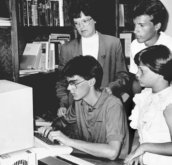A young man uses a 1990s-era computer while others look on.