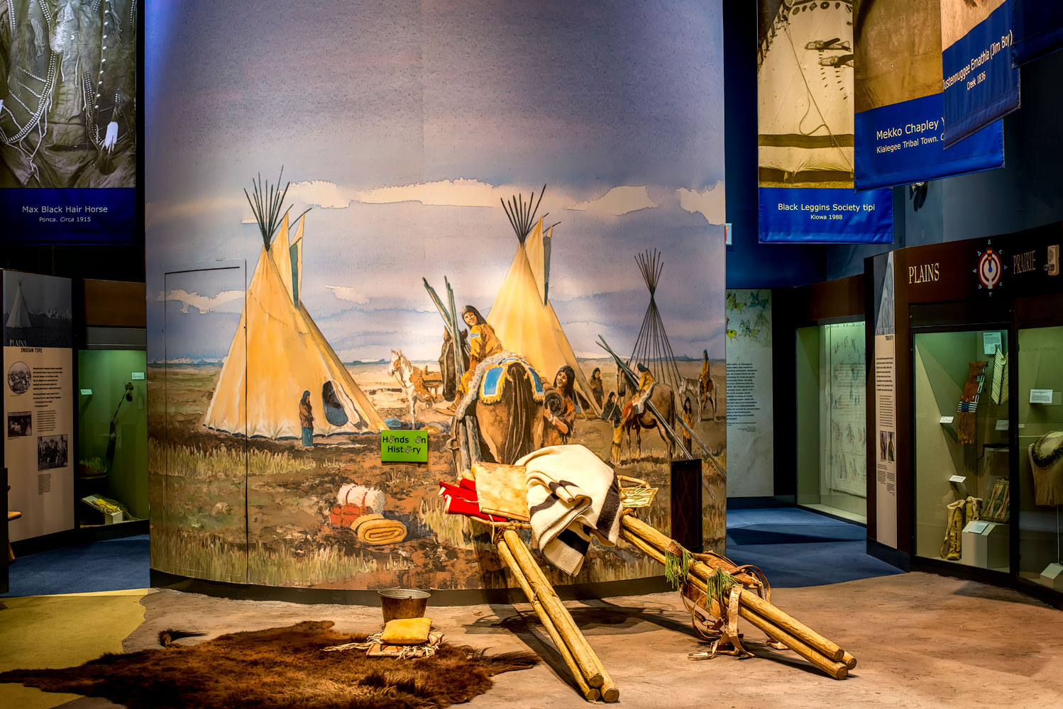 Part of an exhibit with a small tipi mural next to a travois and bison-hide rug