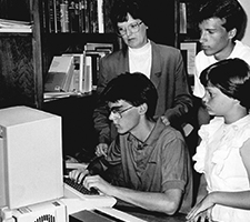 A young man uses a 1990s-era computer while others look on.