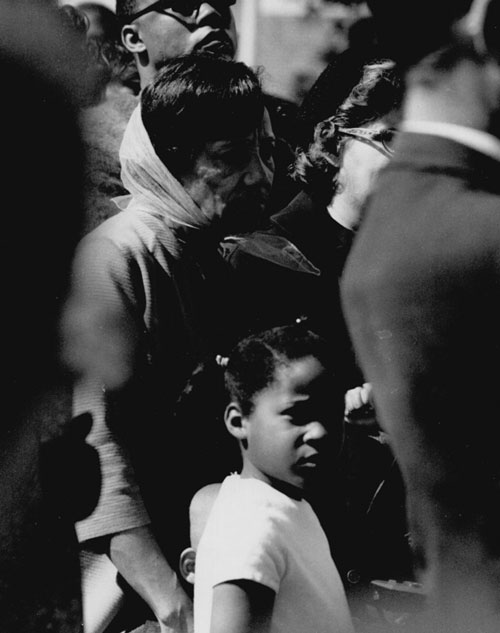 An older Black woman and a young Black child in a crowd of people. The child looks directly at the camera.