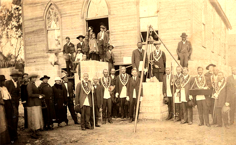 A group of about thirty people, all African American, standing in front of a wooden frame building and stone cornerstones in front of the building. Some of the men are wearing Masonic regalia. 