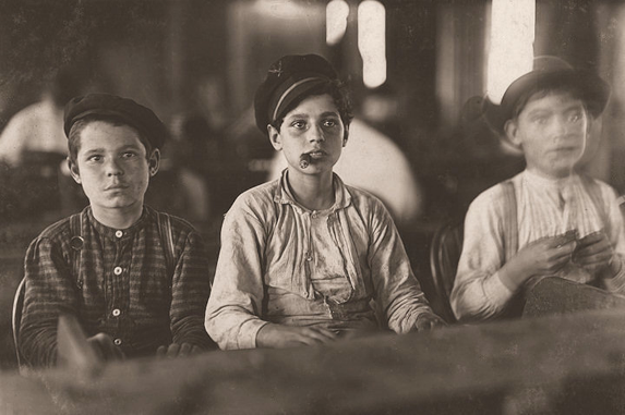 Three young boys make cigars in a factory. One boy holds a cigar in his mouth. Other workers are in the background. 