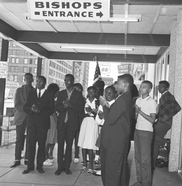 A group of young Black men and women stand together outside a restaurant. Most are dressed in dark suits and dresses. One person is holding the United States flag, and other businesses are visible in the background.