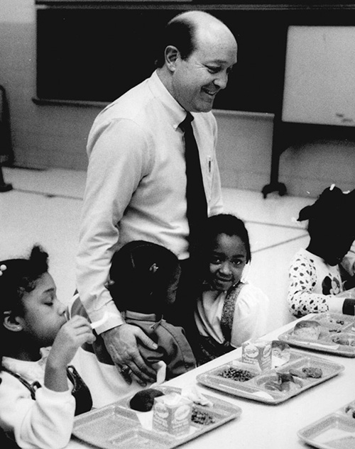 Young girls eat lunch in a cafeteria as the principal pats one on the back.