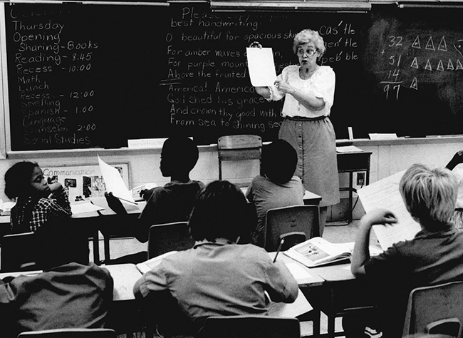 A teacher shows a piece of paper to children seated in desks in front of a chalkboard.